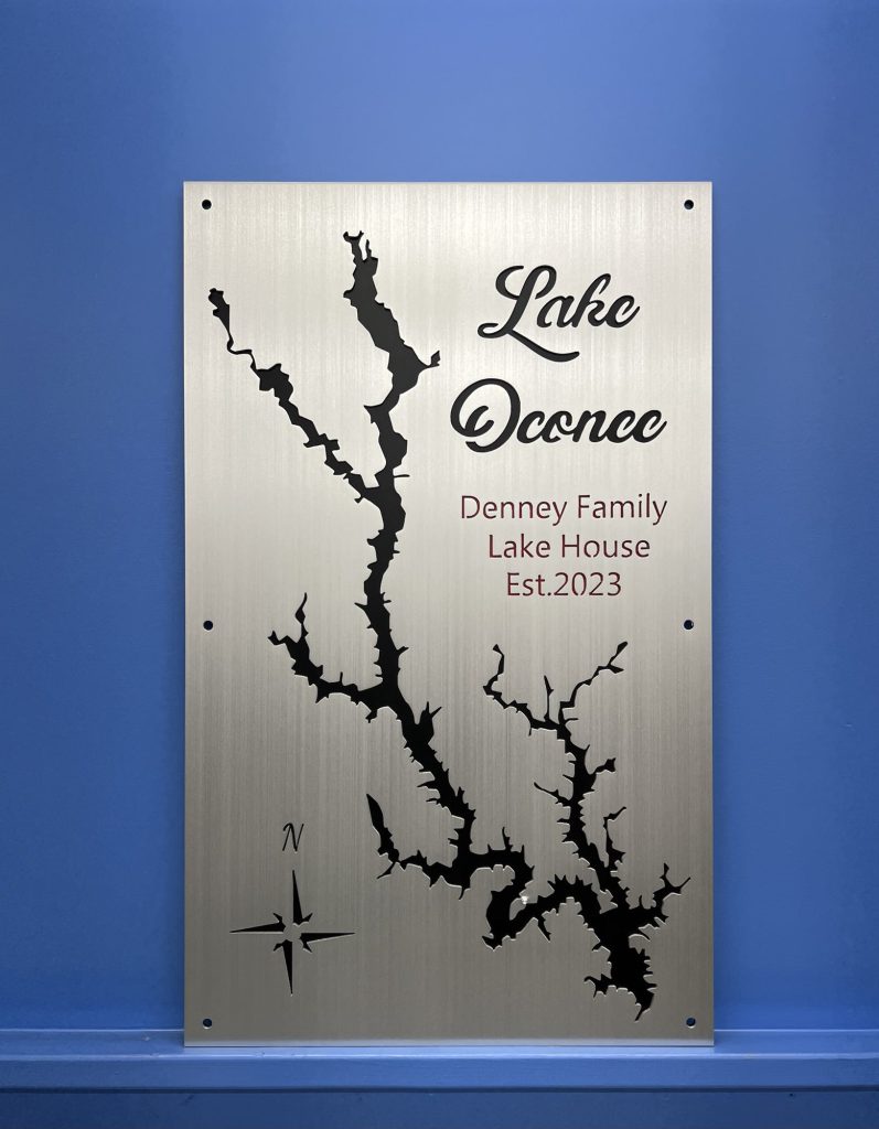 Lake Oconee map on brushed metal, "Denney Family Lake House Est. 2023" text, mounted on a blue wall.