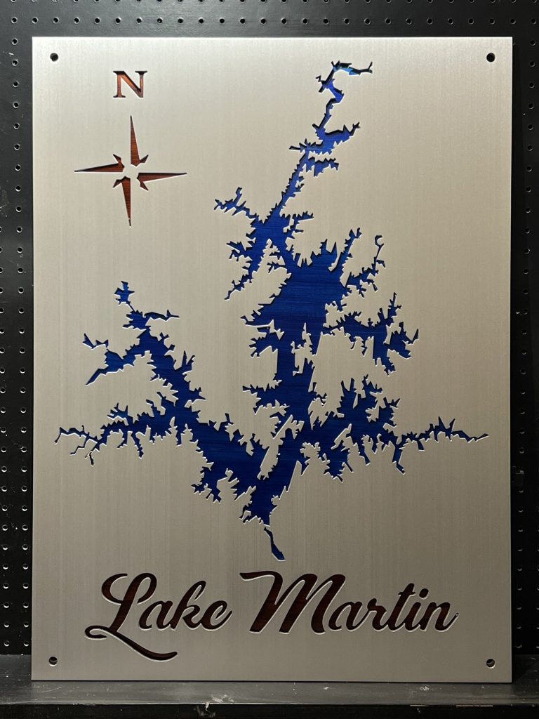 Lake Martin metal map with blue cutout and compass rose on brushed metallic background, mounted on a perforated black surface.